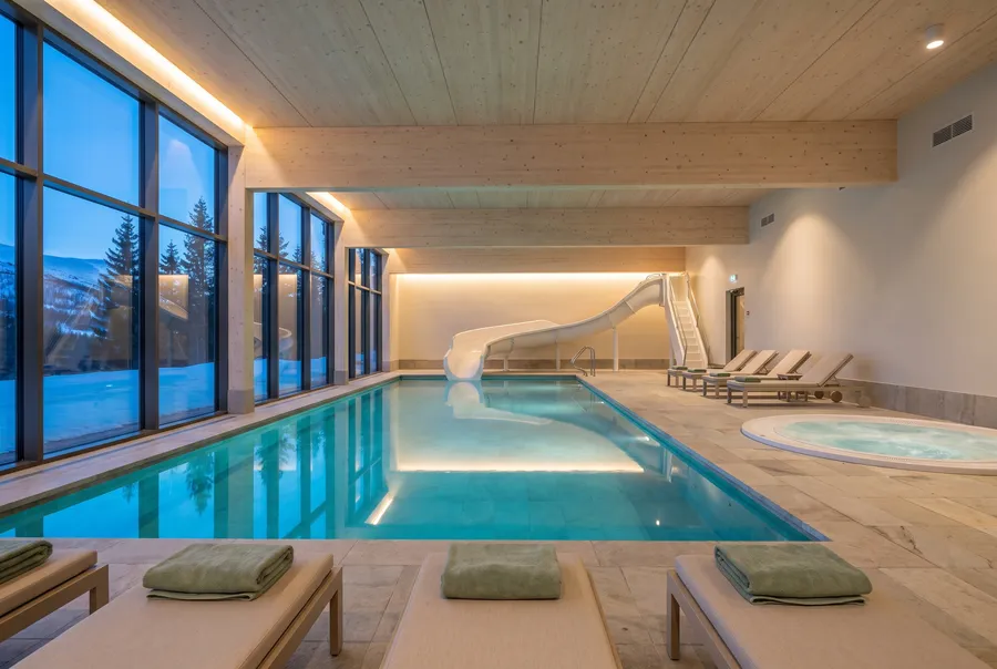 A heated indoor pool with a curving white waterslide and a round hot tub, pale wood beams and a window wall onto snowy spruce at dusk